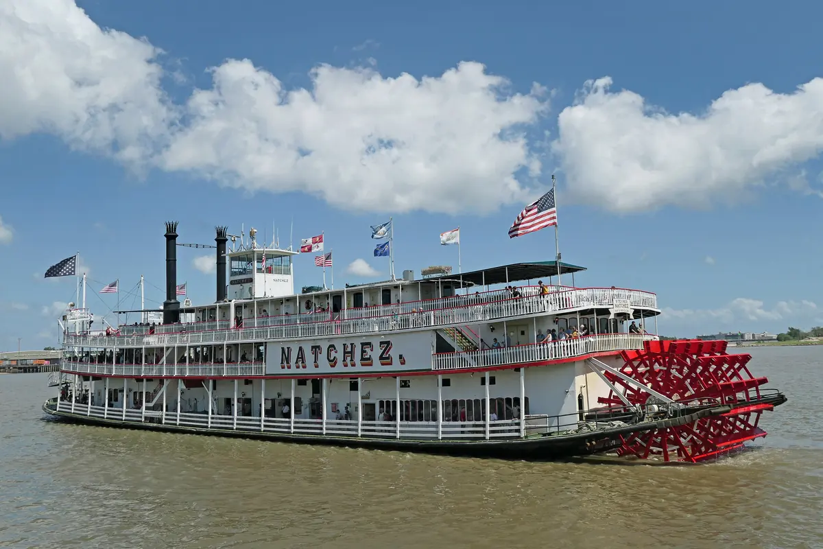 The Steamboat Natchez under way on the Mississippi, a white four-deck sternwheeler with two black chimneys and a red paddle wheel at the back